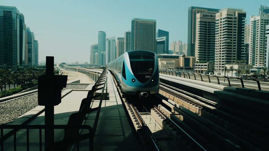 Fotogrāfija, kurā redzams moderns Dubaijas metro vilciens, kas pārvietojas pa paaugstinātu sliežu ceļu.